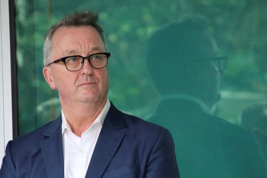 Victorian Health Minister Martin Foley in front of a green-glass window, looking off into the distance.