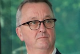 Victorian Health Minister Martin Foley in front of a green-glass window, looking off into the distance.