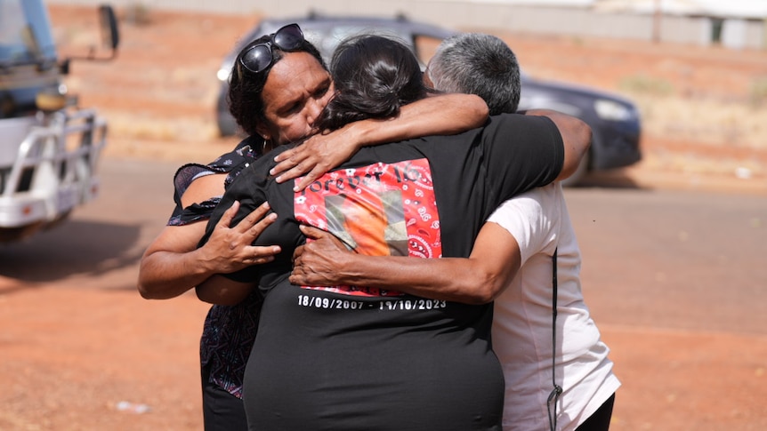Three Indigenous women hug outdoors in front of a small white bus and a car parked in the background.