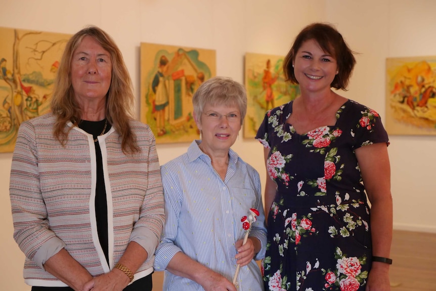 Three women stand inside an art gallery in front of whimsical, 50s style paintings.