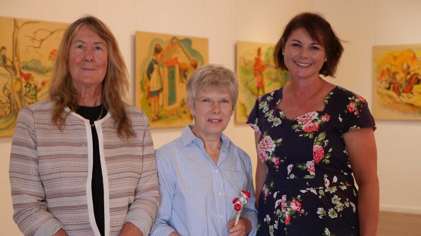 Three women stand inside an art gallery in front of whimsical, 50s style paintings.