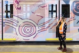 A woman in a high-vis vest stands in front of a painted tram, looking up at the artwork of an Indigenous man's face