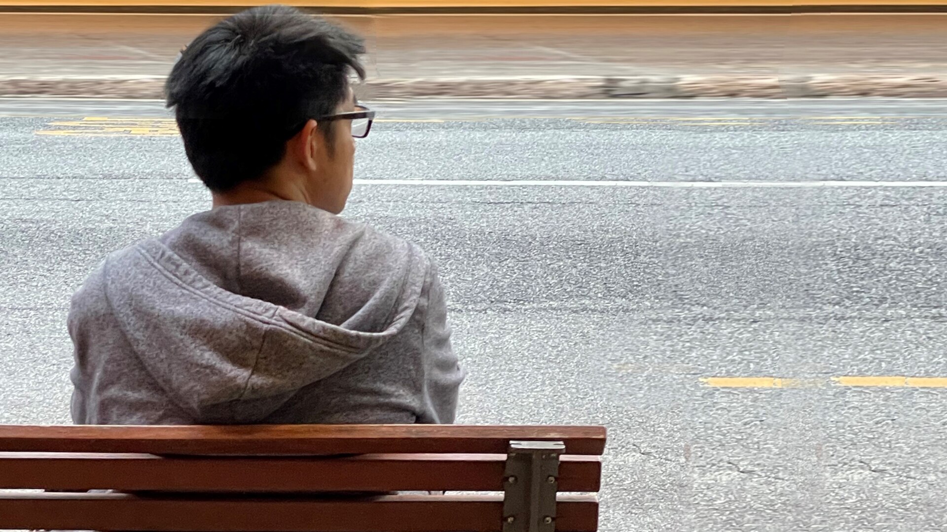 A man with dark hair, glasses and a hoodie sitting alone at a bus stop bench looking to his right.