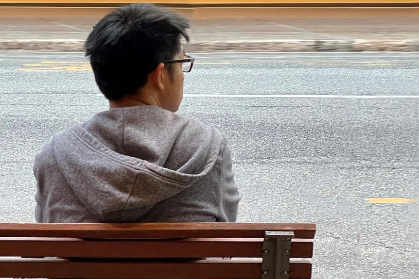 A man with dark hair, glasses and a hoodie sitting alone at a bus stop bench looking to his right.