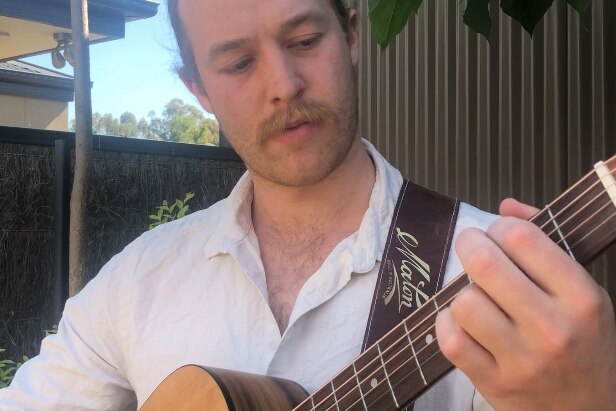 A moustachioed young man wearing a white shirt plays guitar in an outdoor setting.