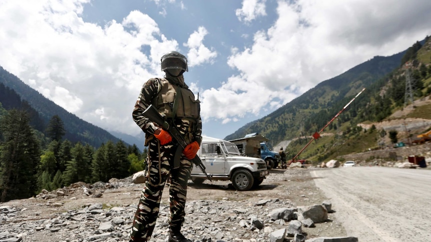 A man in army fatigues stands in front of a jeep and mountains.