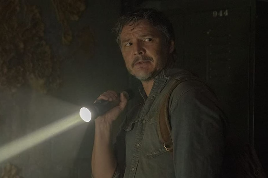 Pedro Pascal as Joel in HBO's The Last of Us holds a flashlight and looks over his shoulder.