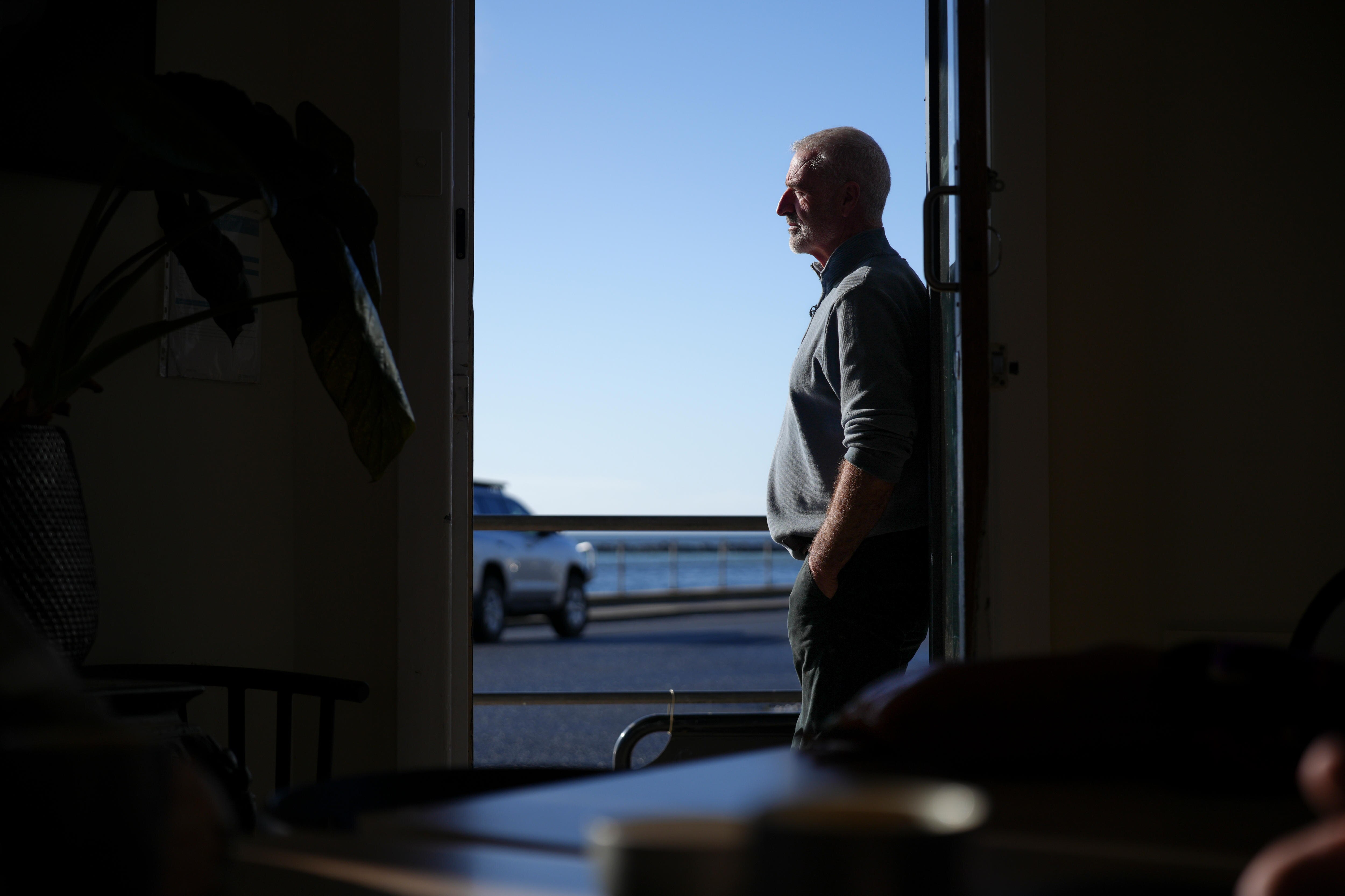 Un hombre se apoya en la puerta de un café, con una cara recta y manos en el bolsillo. Fuera de la puerta hay un paisaje oceánico.