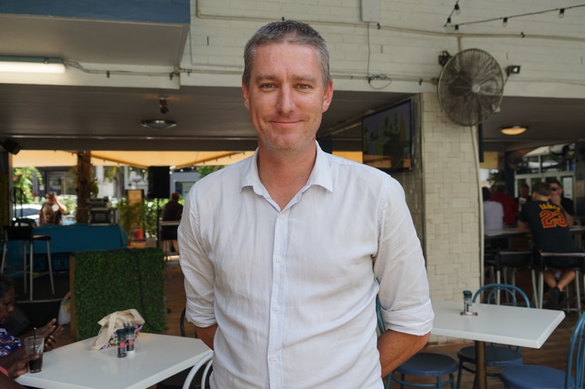 A man in a white shirt outside stares at the camera and smiles.