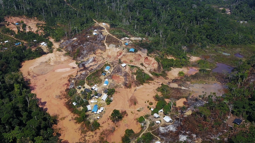 A wide aerial view of cleared land topped with buildings, surrounded by forest