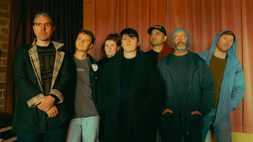 Seven members of Cash Savage and the Last Drinks stand in front of a curtain in a dark room.