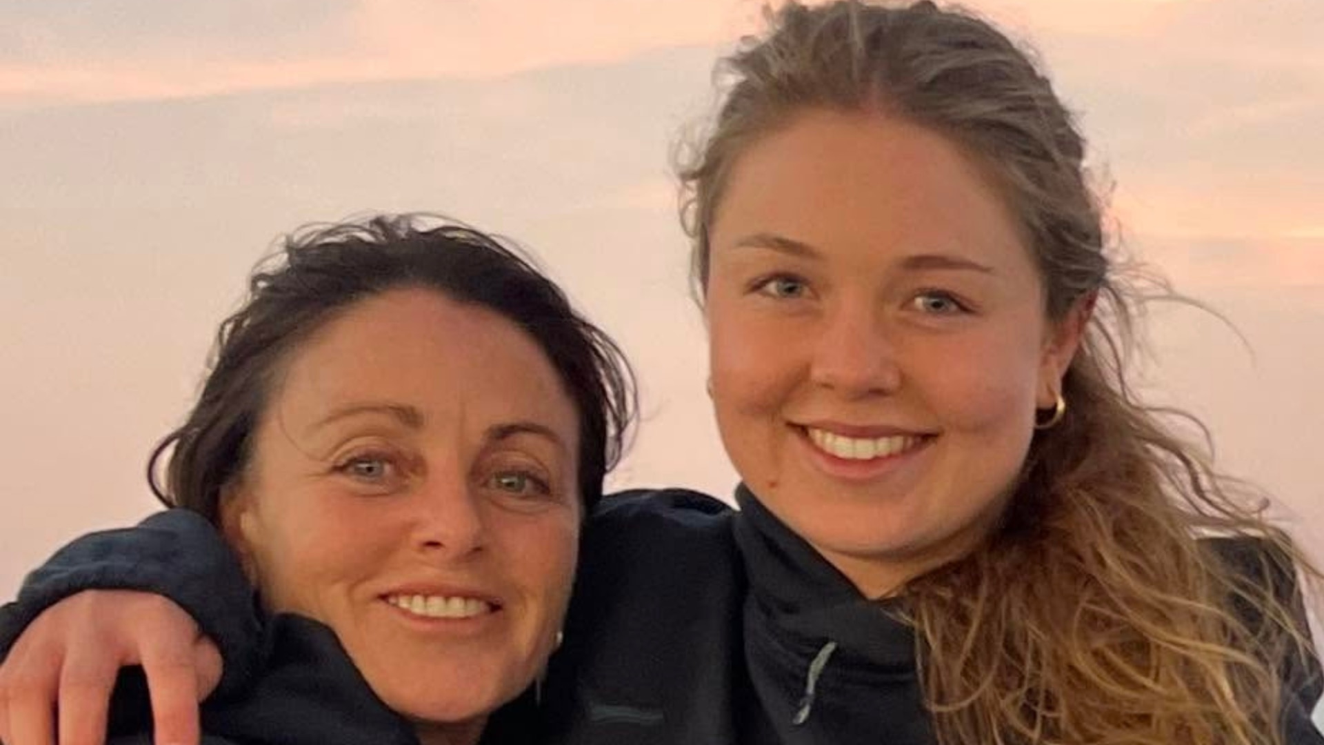 A close up of the faces of two women, they have their arms around one another.