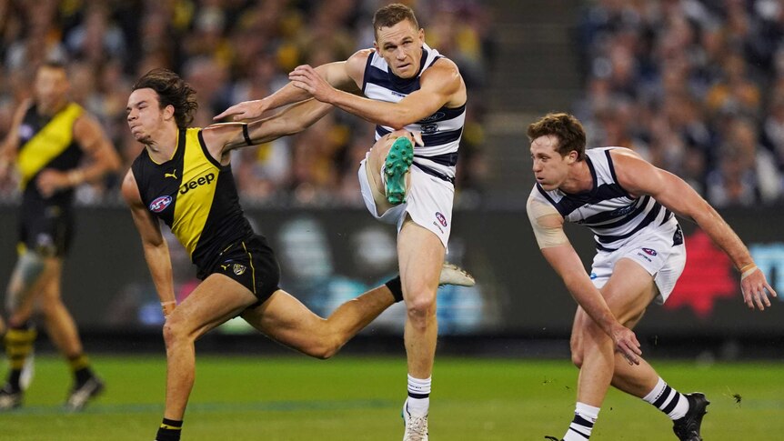 A male AFL player kicks the ball with a teammate and an opponent on each side.