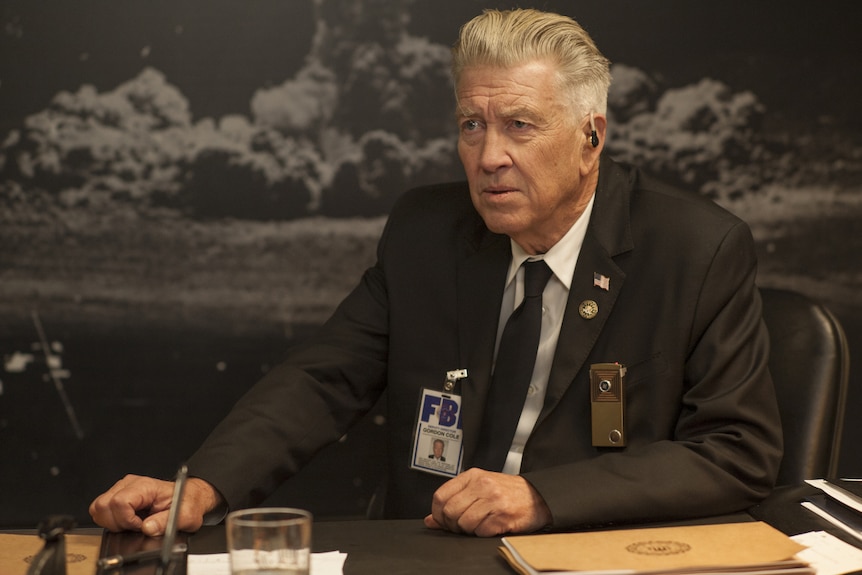 David Lynch, wearing a police uniform and FBI badge, sits at a desk.