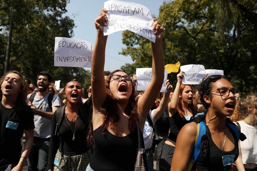 People protest in front of the National Museum holding "Erased our history" and "Education is investment" signs.