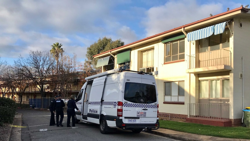 A large white police van is parked in front of a block of units. Three officers stand in front.