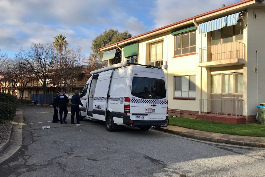 A large white police van is parked in front of a block of units. Three officers stand in front.