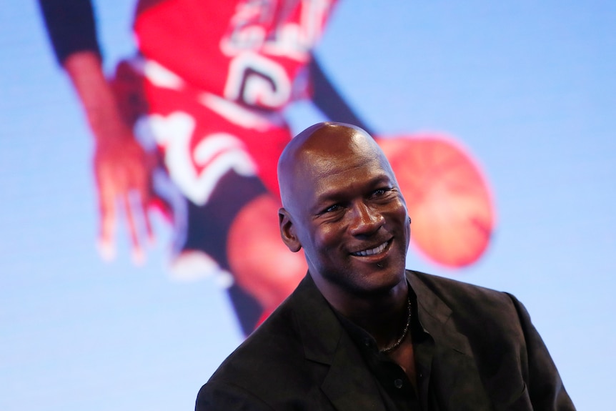 Michael Jordan smiles, sitting in front of a large screen with a picture of him playing for the Chicago Bulls.