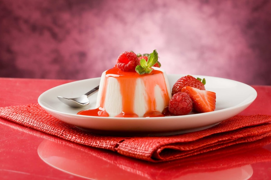 A panna cotta dessert with raspberries and strawberries on a white plate with a silver spoon.