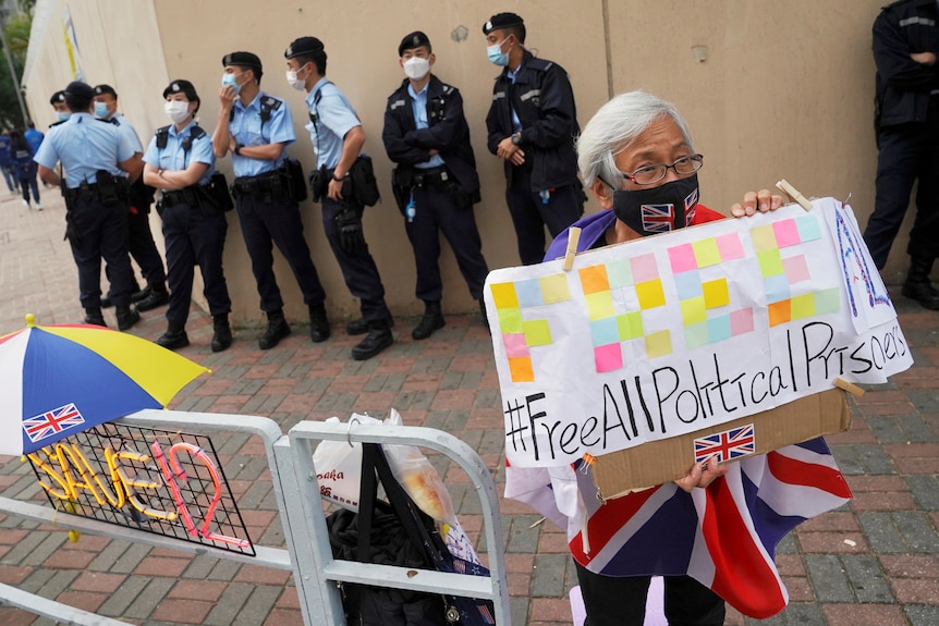 A white-haired man hold a colourful sign in front of a line of police officers in front of a building.