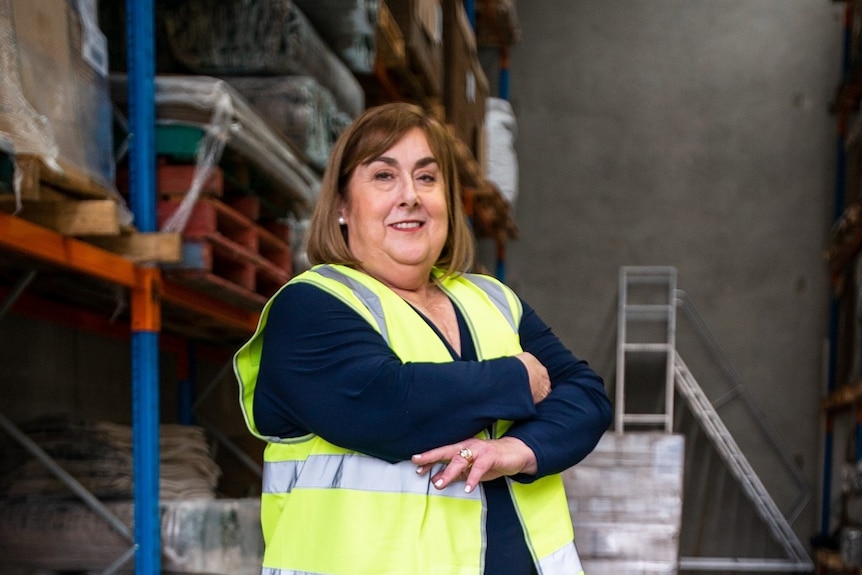 A women in a high visibility vest and arms crossed smiles at the camera. She is standing in a factory.