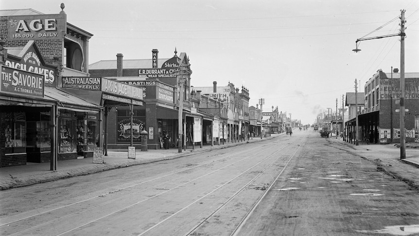 A black and white photograph of a dirt road with tram tracks lined by shops.