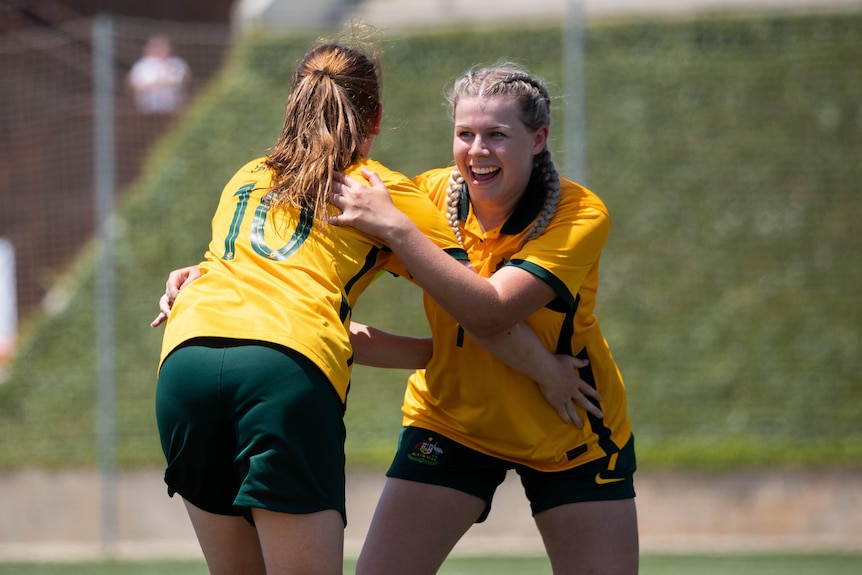 Two female soccer players wearing yellow and green hug after scoring a goal