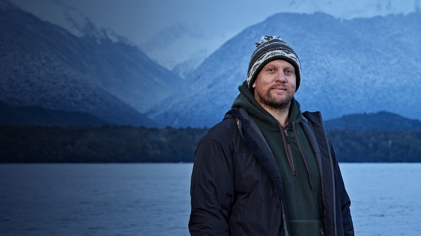 Jason Allwood stands in front of a large lake. There is a snow-capped mountain behind him. 
