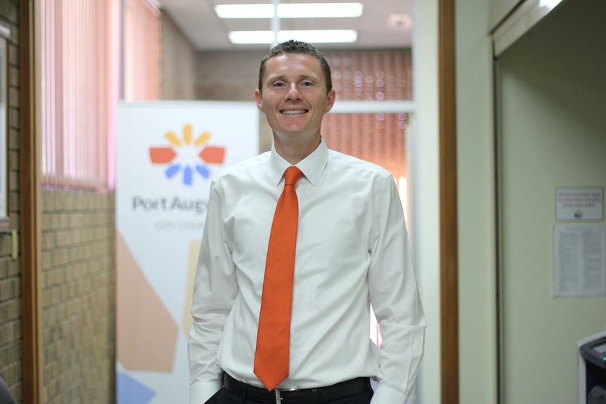 A tall white man with an orange tie smiles at the camera