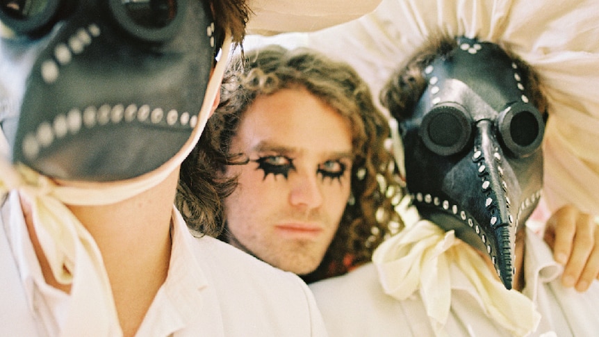 Noah Dillon (curly hair, black eye make up) is stood between two figures wearing plague doctor masks