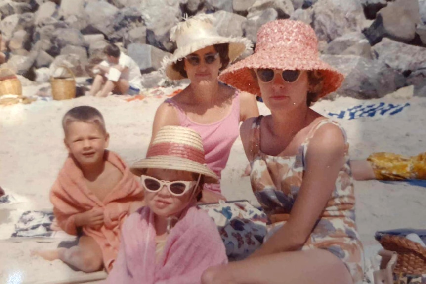 Two women on beach with two kids, wearing colourful hats and bathing suits
