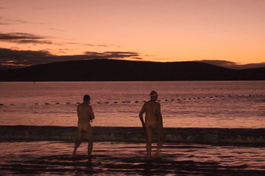 Two men, nude, walk towards the tide as the sun begins to rise.