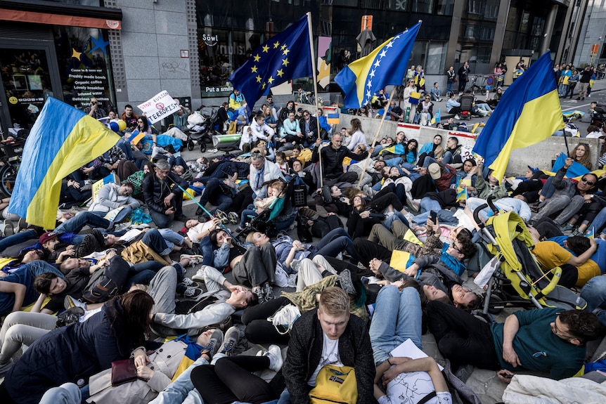 Hundreds of people lie on concrete outside a building. Many hold up Ukrainian and EU flags.