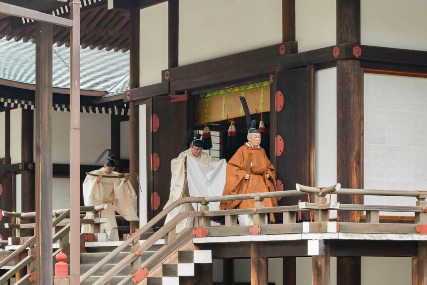 Emperor Akihito in bright orange ceremonial dress walks across a traditional Japanese balcony as two servants trail him.
