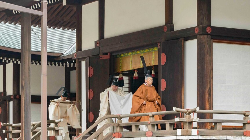 Emperor Akihito in bright orange ceremonial dress walks across a traditional Japanese balcony as two servants trail him.