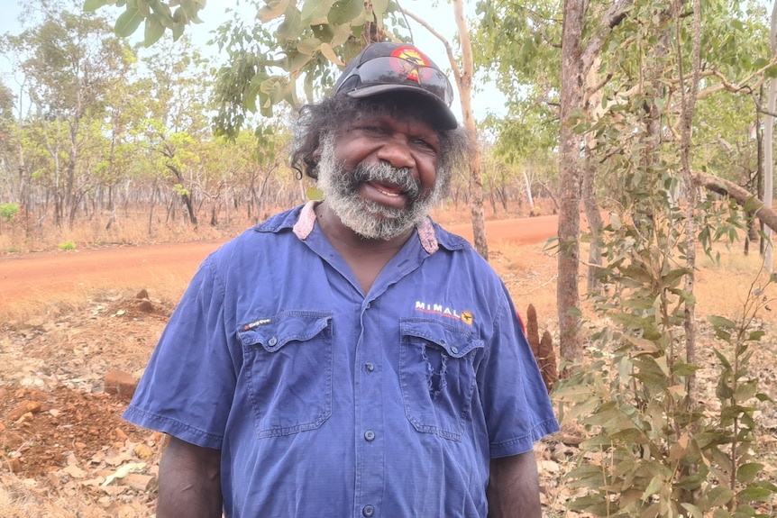 A ranger, wearing a blue uniform and a black cap stands in native bushland and is smiling to the camera.