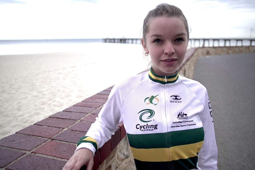 A young woman in cycling jacket stands in front of a beach.