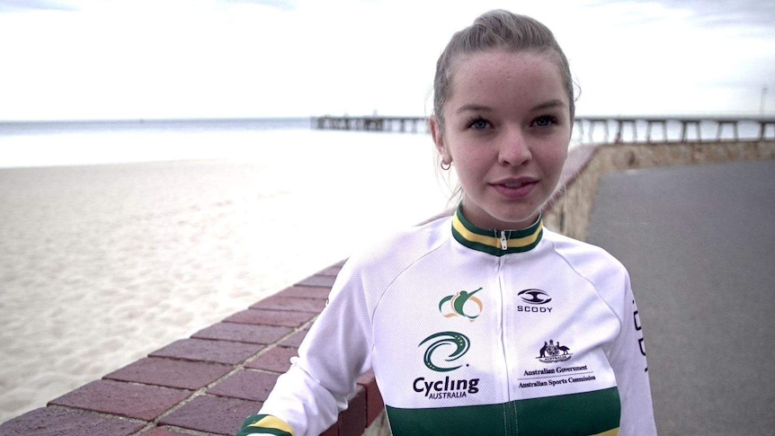 A young woman in cycling jacket stands in front of a beach.