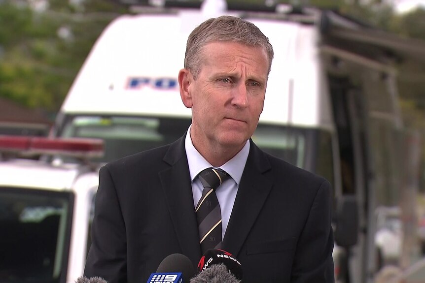 a detective wearing a suit and tie speaking into media microphones at a crime scene with police vans in the background