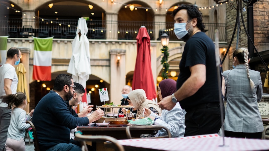 a man standing next to a table of diners wearing a mask 