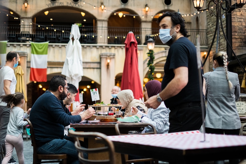 a man standing next to a table of diners wearing a mask 