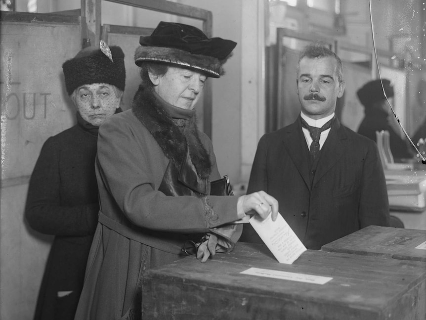 A woman with a large hat dressed in early 20th century fashion looks down as she puts her ballot in a box.