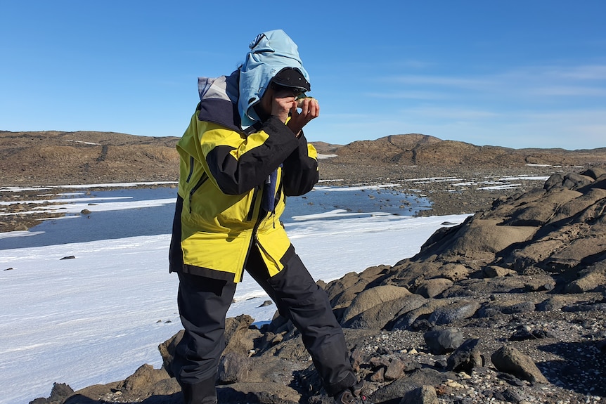 A woman in a yellow and black jacket peers through a small microscope while standing on a rocky outcrop amongst snow. 