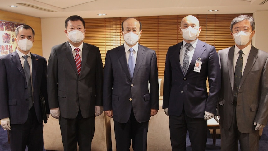 Five men wearing masks and suits pose for a photo inside an office.