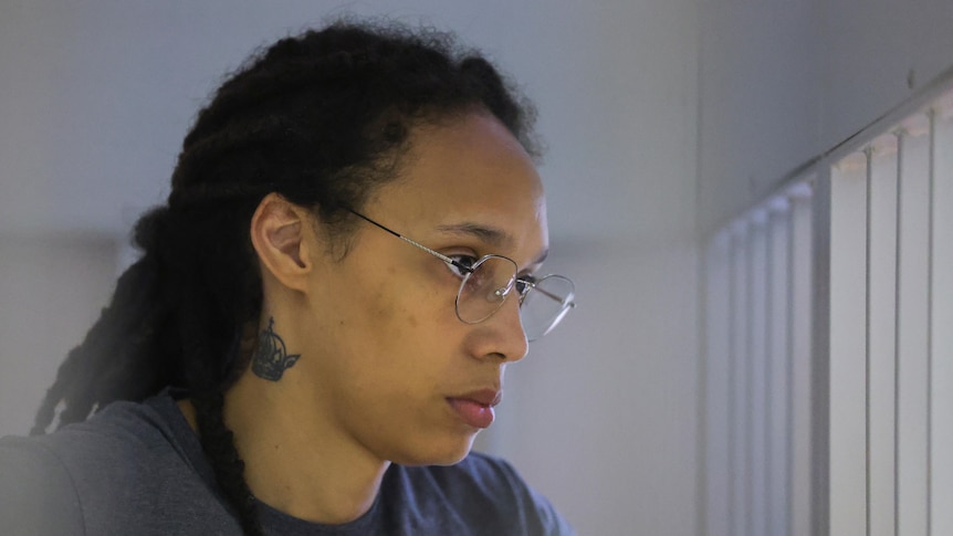 A close-up of a solemn Black woman wearing wire-framed glasses and grey T-shirt