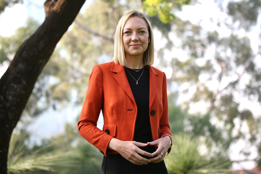 A mid-shot of Mia Davies wearing a red jacket and black shirt posing for a photo outdoors with her hands poised together.