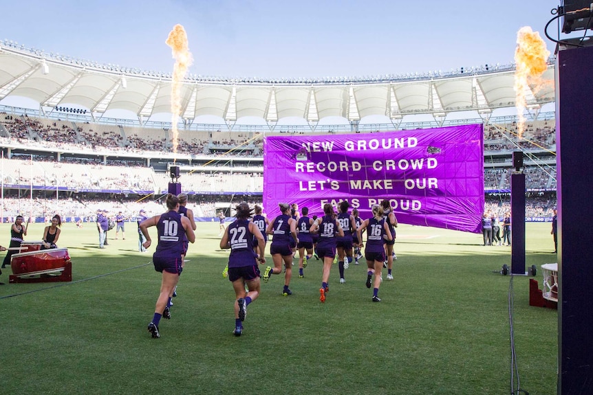 The Fremantle Dockers AFLW team runs on to Perth Stadium and towards a purple banner.