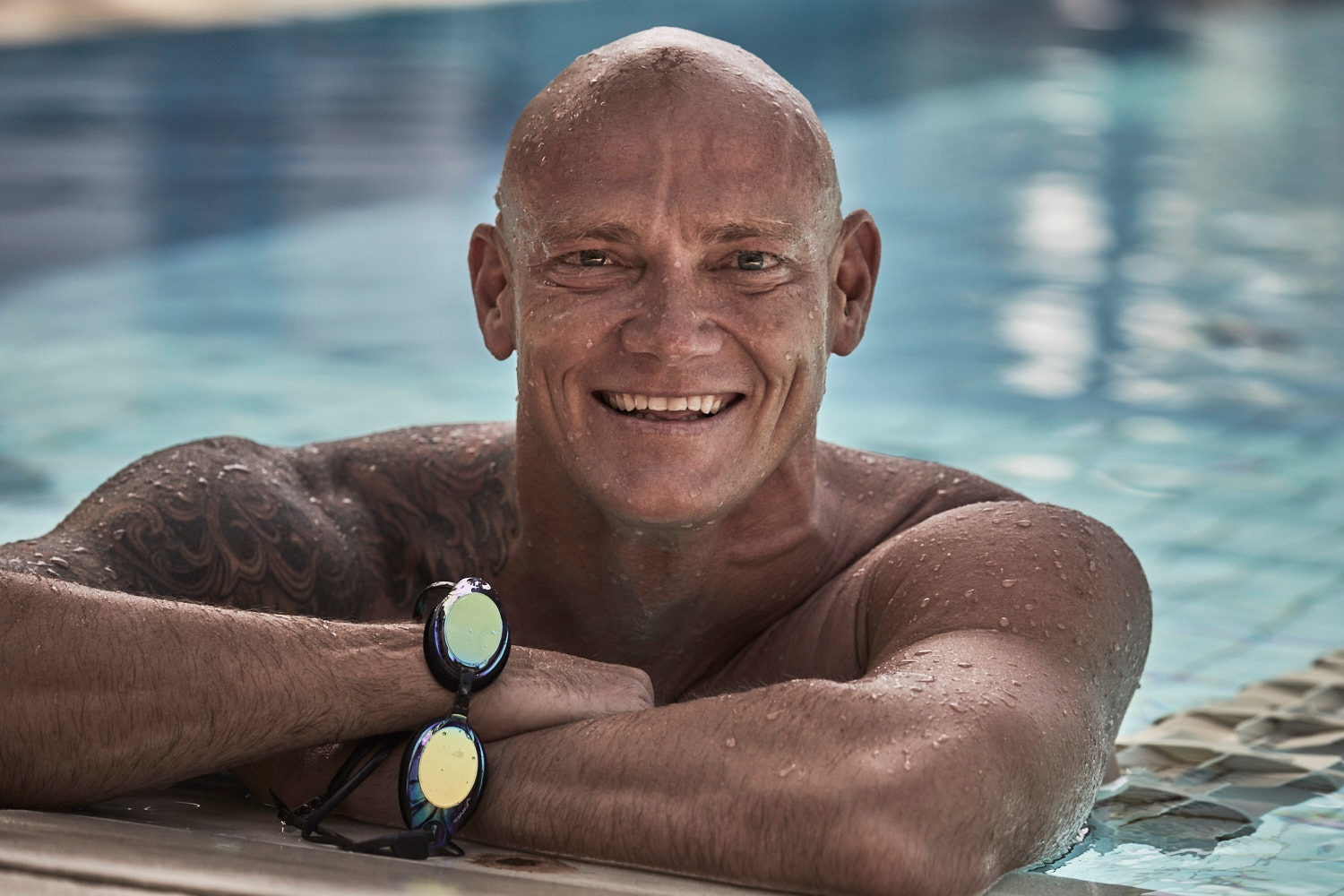 Bald Michael Klim, shirtless, leaning on the edge of a pool. He has water droplets on his skin and is smiling