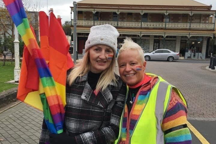 Two women with rainbows painted on their faces, holding rainbow flags, standing in front of a historic building