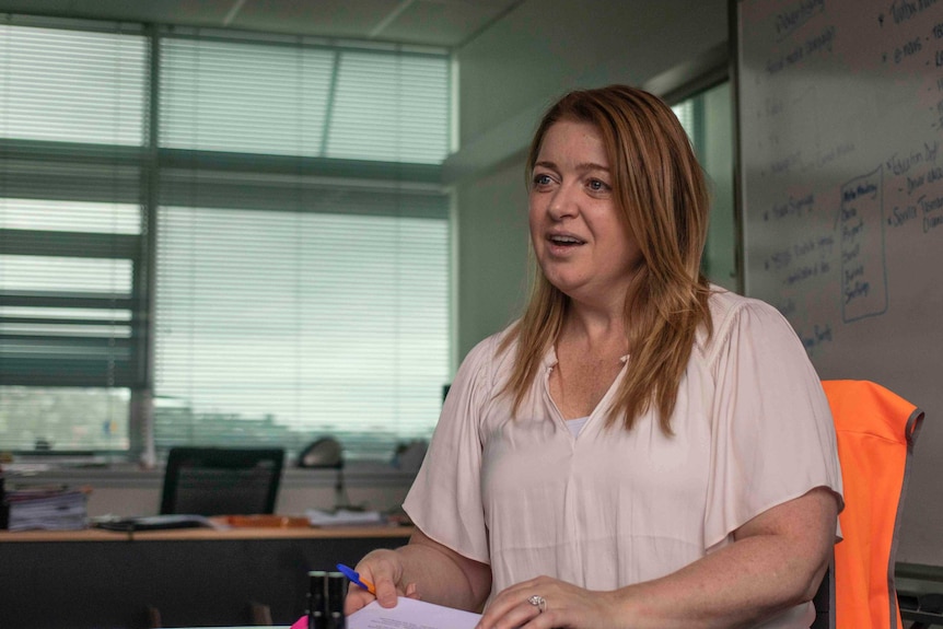 A woman with brown hair sits in front of a whiteboard